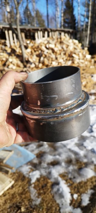 En hand håller en brun och silverfärgad metallkomponent, bakgrund med vedhög och smältande snö.