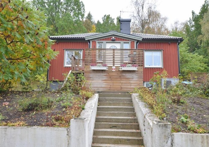 Ett rött trähus med veranda, trädgårdstrappa, fönsterblommor och höstlöv.