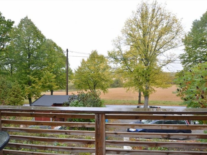 Utsikt från en balkong med träd, ett fält, en bil och ett litet hus. Höstiga färger och fuktig atmosfär.