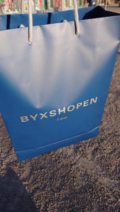 En blå shoppingväska med texten "BYXSHOPEN Falun" hänger i solljus, reflektioner på trottoarens yta.