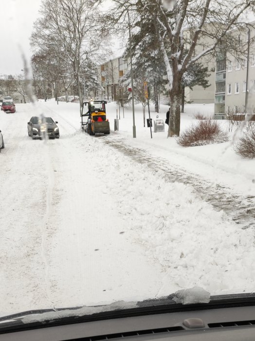 Snötäckt gata med lastmaskin, bilar och flerbostadshus, sedd från bilens insida.