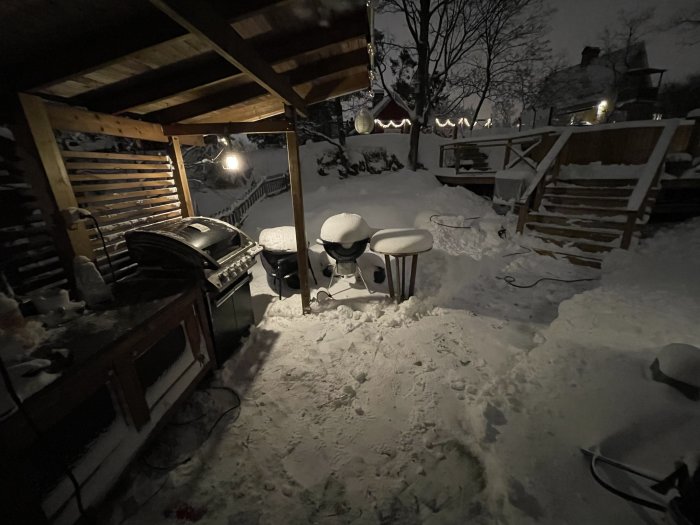 Uteplats täckt av snö vid natt, grill, möbler, ljusslingor, trätrappa, vintrig trädgårdsmiljö.