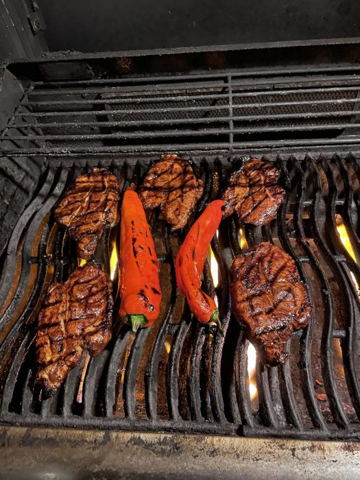 Grillade biffar och röda paprikor på en gasolgrill med tydliga grillränder och synliga lågor.