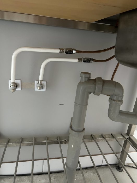 Vattenrör och avloppsrör under diskbänk, fastsatt vid vägg, struktur med metallhylla nedanför.
