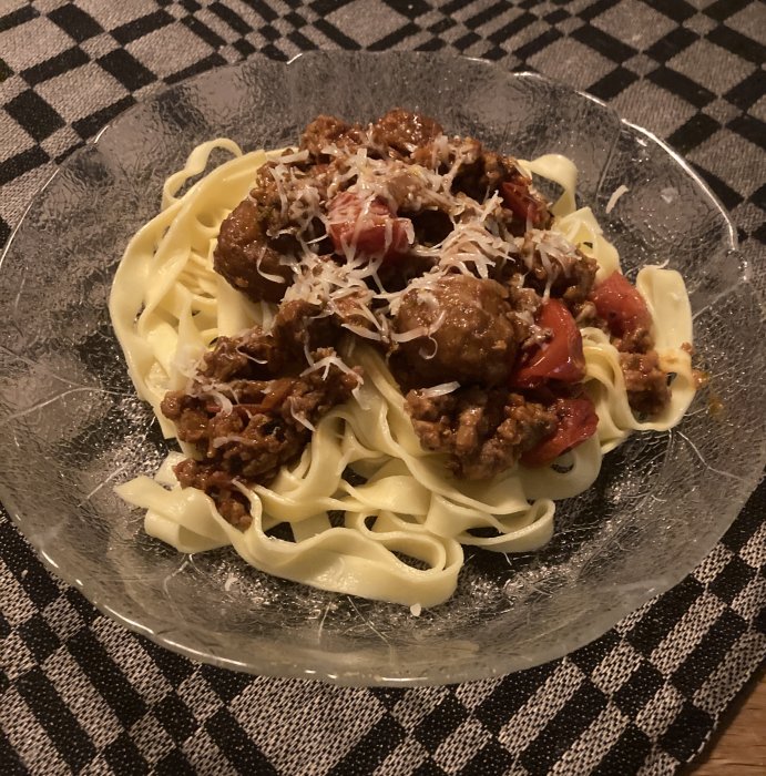 En tallrik med pasta, köttbullar, tomatsås och riven ost på rutig duk.