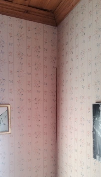 Ett hörn av ett rum med blommig rosa tapet, träpaneltak, och två inramade bilder.