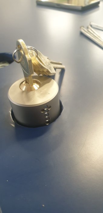 Rund hänglås med nycklar instuckna, på blå yta, ljusreflektion i bakgrunden, fokus på lås och nycklar.