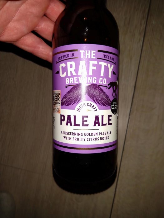 En hand håller en flaska med Crafty Brewing Co. Pale Ale, bryggd i Irland, med citrustoner.