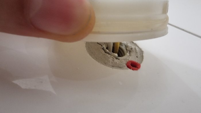 En hand håller en plastdel över ett hål i en vit yta, rör och röd tätning syns.