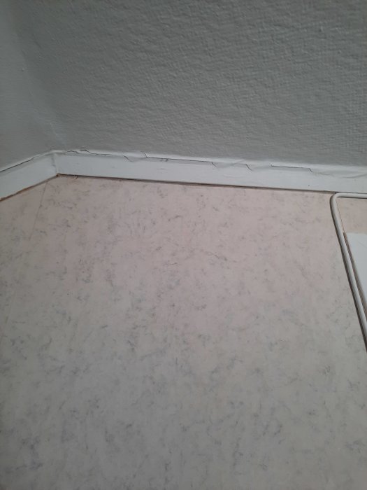 Hörn av ett rum med spruckna vita lister och marmormönstrat golv. Behov av underhåll synligt.