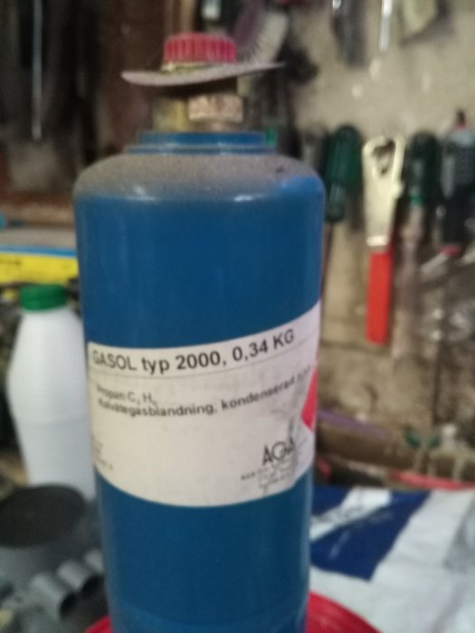 Blå gasolflaska med etikett, verkstadsmiljö i bakgrunden, innehåller gasblandning, AGA-märkt, 0,34 KG.