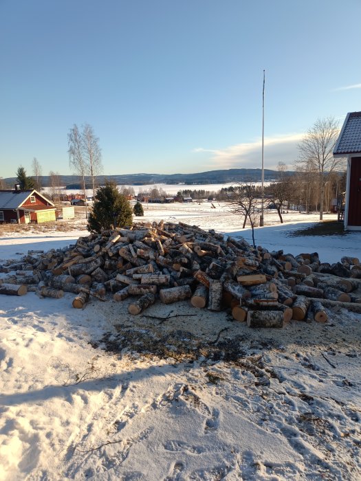 En hög med ved framför röd stuga, snö, blå himmel och en flaggstång på en klar vinterdag.