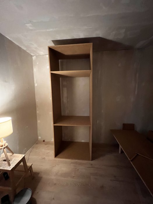 Ommöblerat rum under renovering med tom bokhylla, stegpall och golvlampa.