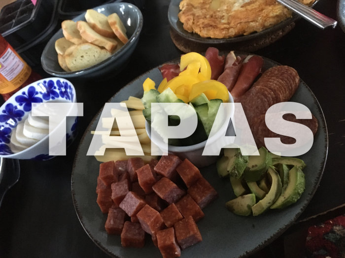 Variation av tapas: charkuterier, ost, bröd, avokado, paprika. Texten "TAPAS" täcker delar av bilden.