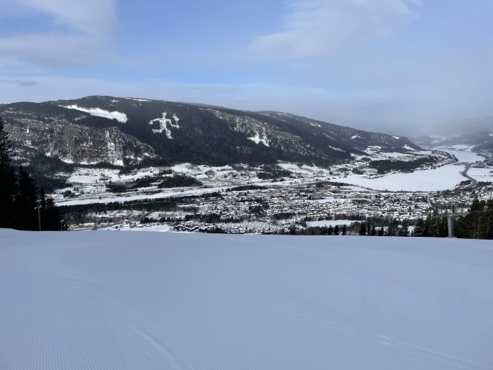 Vinterlandskap med snötäckt stad, berg, skidbackar och frusen sjö under blå himmel.