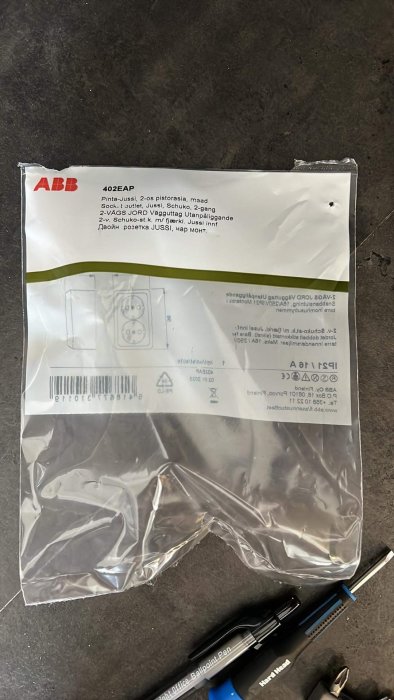 Förpackning ABB, vägguttag, beskrivning, streckkod. Pennor, skruvmejsel synliga på mörk ytter.