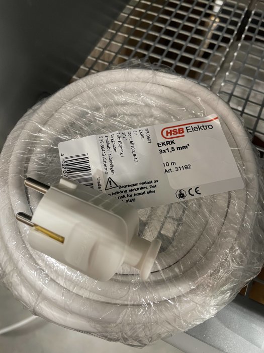 Elkabel på rulle med etikett och en vit stickkontakt framför, ligger på metallhylla.