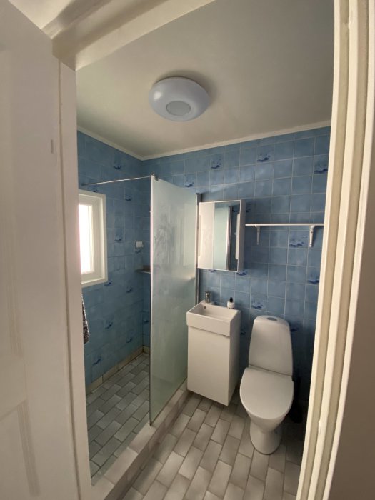 Modernt badrum med blå kakel, duschhörna, toalett, handfat, spegel och grått golv.