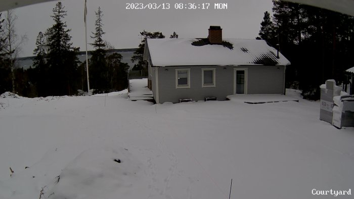 Ett grått hus med snötäckt tak och omgivning, träd i bakgrunden, molnigt, gryning eller skymning.