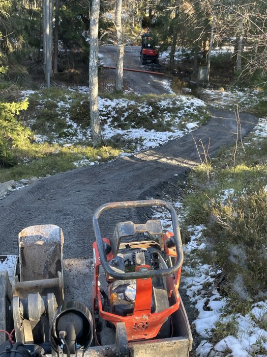 Minigrävare förgrunden, terräng, skog, snötäckt mark, och vattenpöl synlig. Maskin på väg i bakgrunden.