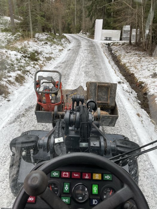 Vy från traktorhytten, snöig skogsväg, släpfordon bakom, vinterdag, instrumentbräda, grå himmel.