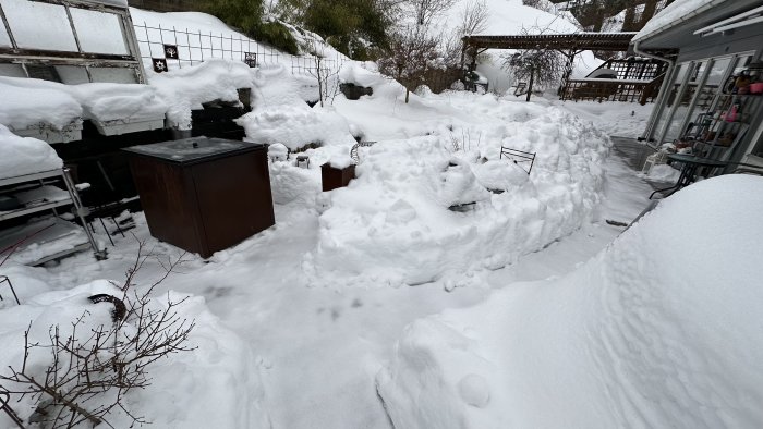 Utomhusområde täckt av tjockt snötäcke, trädgårdsmöbler och grillparti synliga, kallt vinterlandskap.