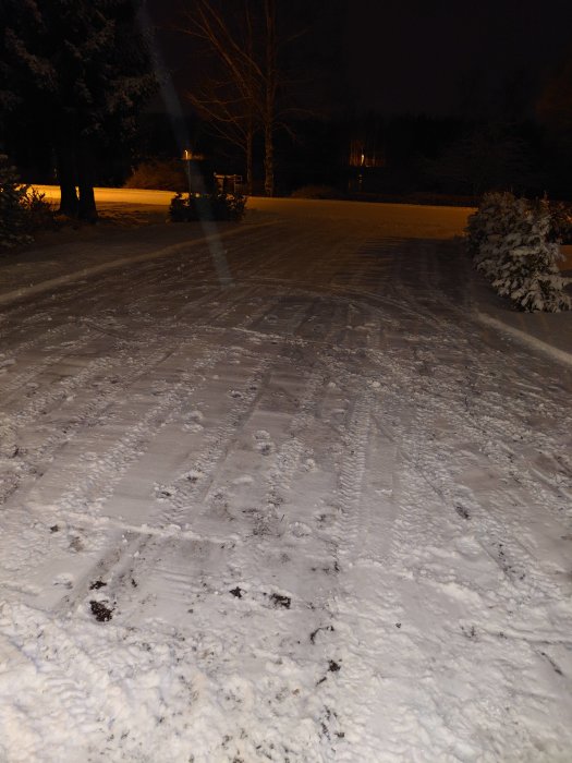 Natt, snö täcker marken och vägar, fotspår och bilspår synliga, mörk himmel, träd.