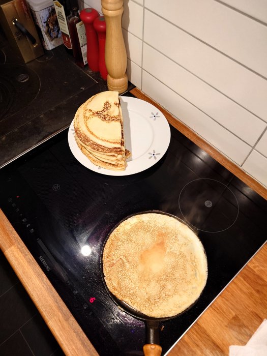 Pannkakor tillagas i stekpanna, staplade på tallrik, kökstillbehör i bakgrunden, spishäll.