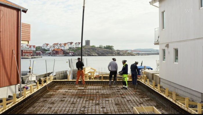 Byggplats vid vatten med arbetare, hus, brygga och havsutsikt i molnigt väder.