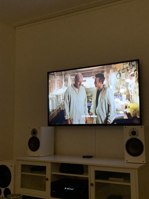 Två personer på tv-skärm, interiör, hemmabio, högtalare, undertexter, vardagsrum, kvällstid.