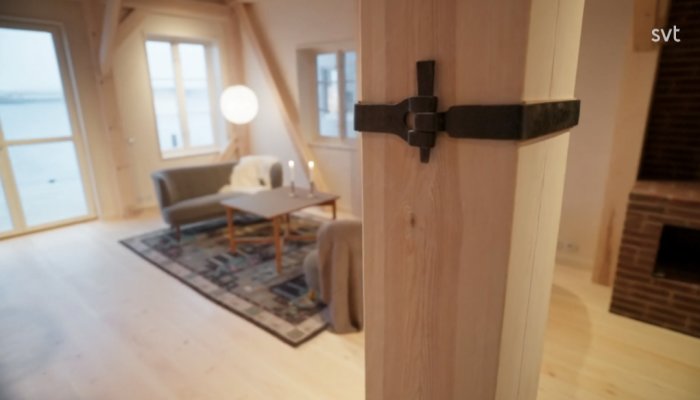 Ljust vardagsrum med havsutsikt, trägolv, soffa, matta, och lampa i skymning, förgrunden visar en dörrkarmdetalj.