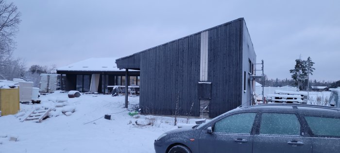 Modern träbyggnad under konstruktion, omgiven av snö, byggmaterial på marken, skymning, parkerad bil framför.