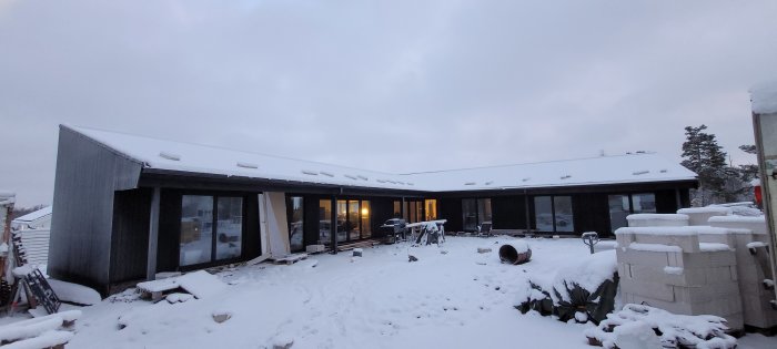 Modernt hus med stora fönster, snö, nedgång sol, trädgårdsmöbler, grill, lugn vinterkvällsstämning.