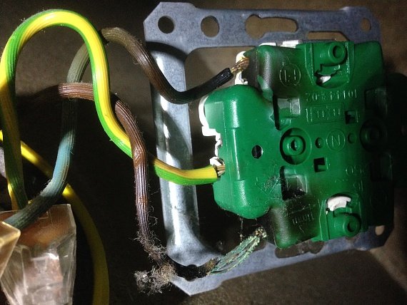 Eluttag med grönt hölje, omslutet av elektriska kablar, delvis exponerat, monterat på en metallram. Slitage synligt.