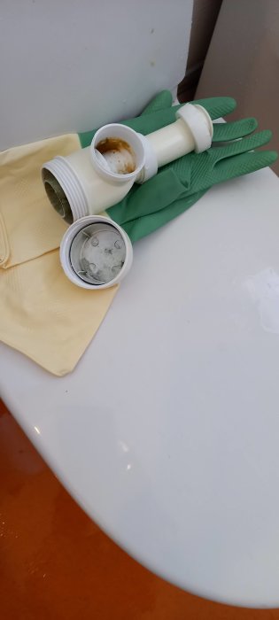 Ett par gröna gummihandskar och delar av ett avloppsrör på en vit yta, potentiellt städ- eller reparationssituation.