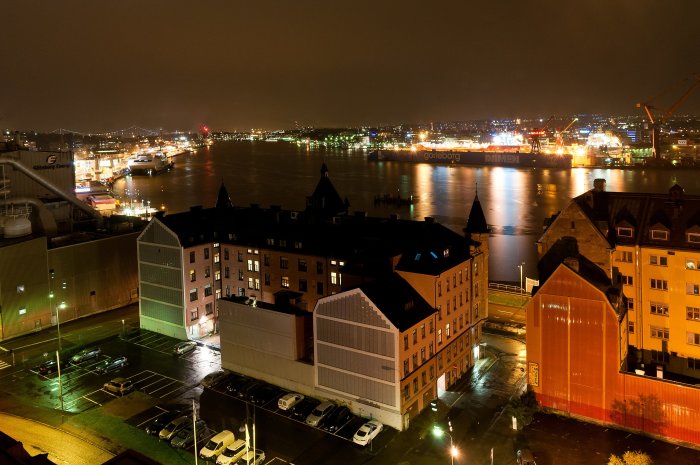 Nattvy över stad med byggnader, belyst hamn, parkerade bilar och reflekterande vattenspegel.
