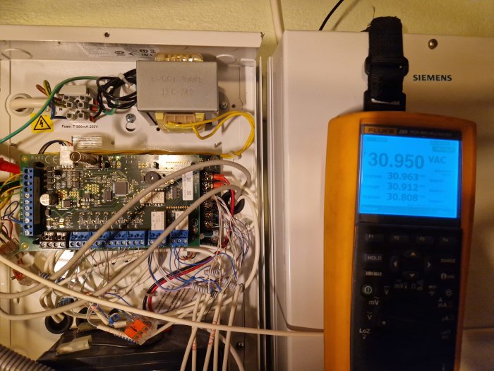 Elektriskt skåp med kablar och elektronikkort, bredvid en multimeter som visar växelspänningsmätningar.