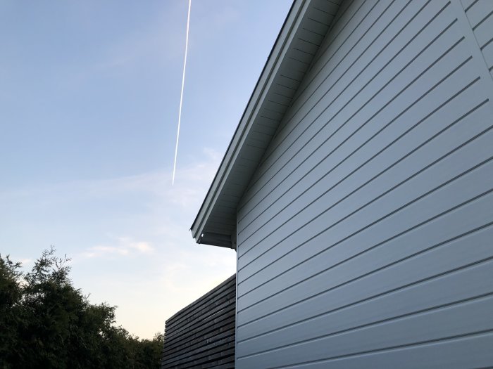 Ett hus med plastfasad, trädetaljer, flygplansspår på himlen vid skymningen.