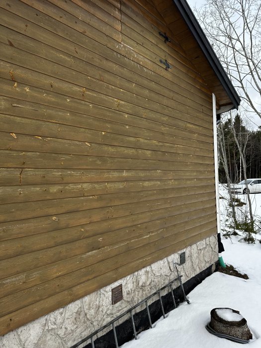 Trävägg på byggnad, snö, inga fönster, dimmig himmel, tre svarta ljushållare, luftventil, avsågad trädstubbe.