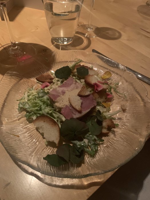 En måltid med sallad, kött, krutonger på guldfärgad tallrik, vattenglas och vinglas ovanpå ett träbord.