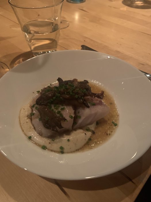 Ett välpreparerat maträtt på vit tallrik med fisk, sås, svamp och strödd gräslök, serveras vid ett träbord.