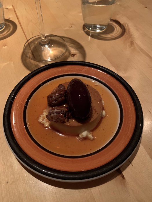 Tallrik med mat, dessert med päron, glass, chokladsås, en vinglasfot och vatten, på träbord.