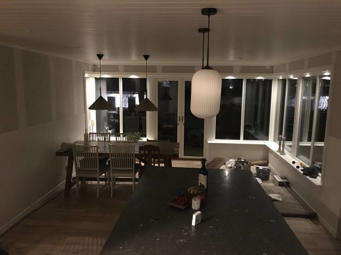 Ett kök på natten med bordslampa, spis, matbord, stolar och fönster. Oordnat och bebott utseende.