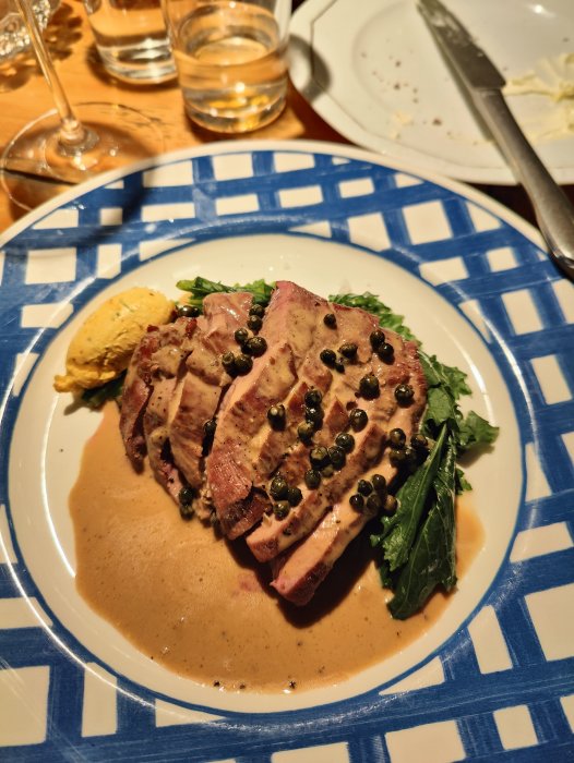 Tonfisksteak med kapris och sås på mönstrad tallrik, delvis uppäten mat, tom tallrik och vattenglas i bakgrunden.