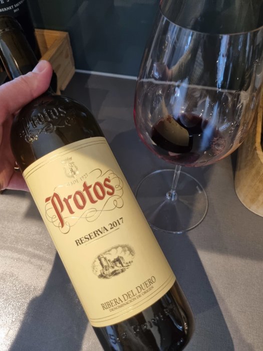 Vinflaska märkt "Protos Reserva 2017" hålls bredvid vinglas med rödvin; Ribera del Duero.