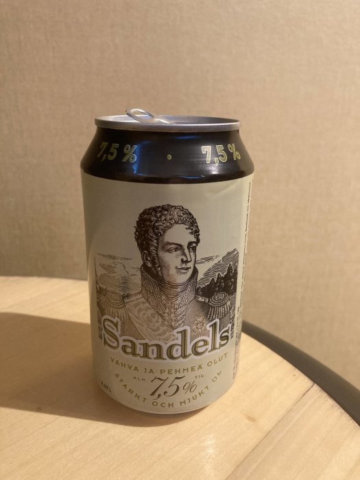 En ölburk med texten "Sandels" och grafik, 7,5% alkoholstyrka, på en träyta mot beige bakgrund.