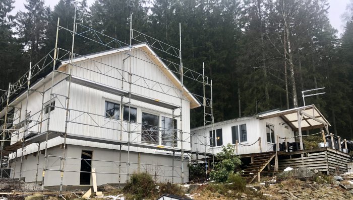 Hus under renovering omgivet av ställningar med skog i bakgrunden och lätt snötäcke.