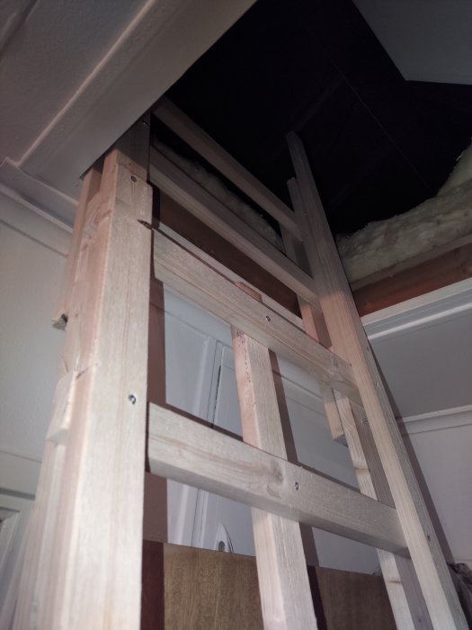 Trästege som leder upp till en öppen vindslucka i ett hus, synligt isoleringsmaterial, snedtak.