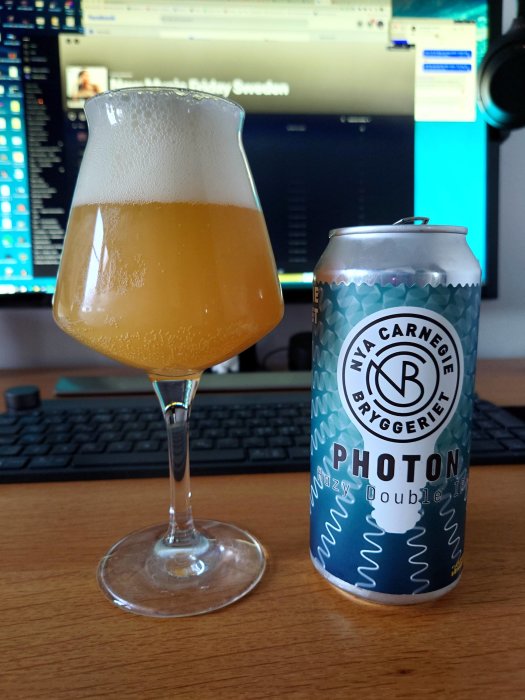 Ölglas med skum på skrivbord framför datorskärm, burk märkt "Photon Hazy Double IPA" vid sidan.