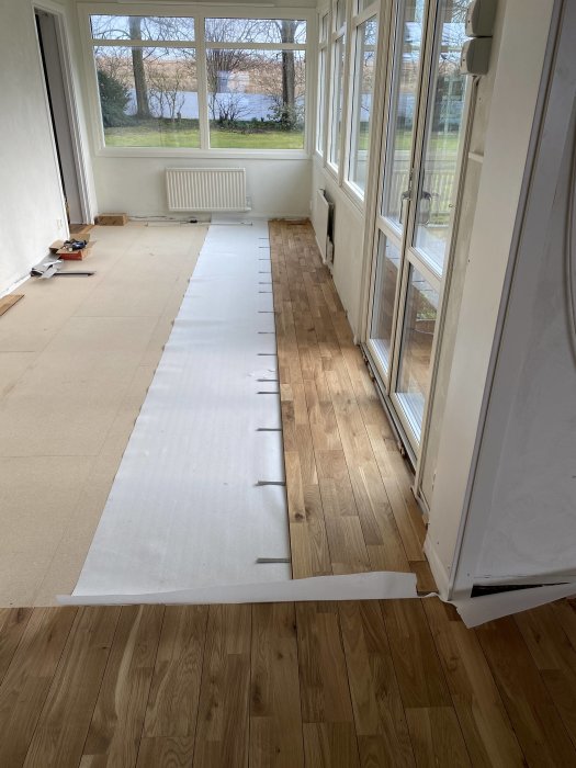 Rum under renovering med trägolv, delvis täckt med skyddspapp, stora fönster med utsikt, ljust och öppet.